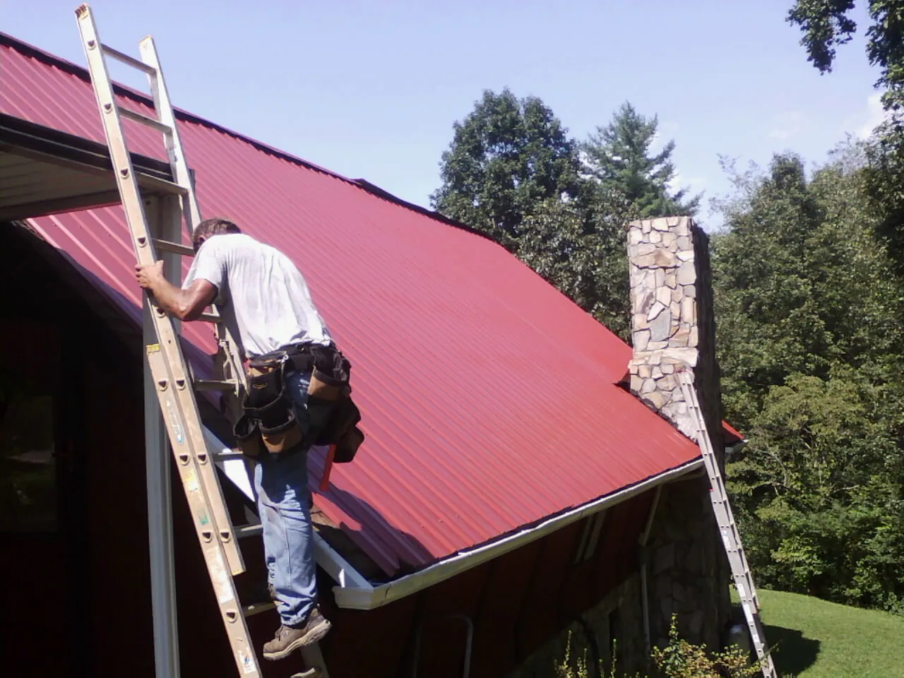 Roofer on a ladder beside a red metal roof and stone chimney — Soffit & Fascia Repair by Roofing Brothers in Levittown